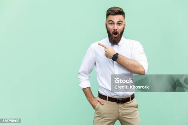 Handsome Young Adult Man With Beard In Shoked Pointing Away While Standing Isolated On Light Green Background Stock Photo - Download Image Now