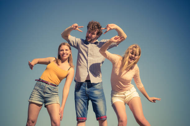 Friendship happiness summer holidays concept. Group of friends boy two girls having fun outdoor stretching arms celebrating, against sky
