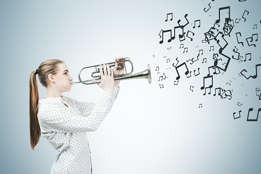Side view of a blonde woman with a ponytail playing a saxophone. Gray wall background with music notes on it.