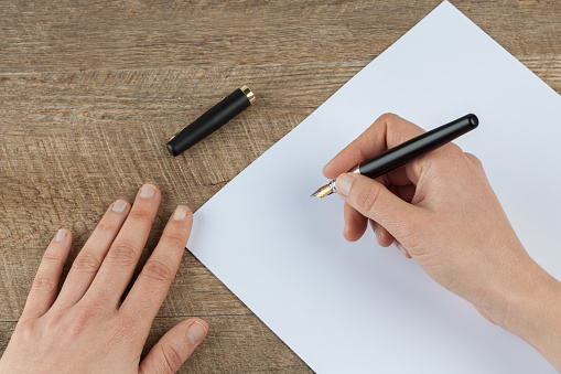 Female hands holding fountain pen and blank piece of paper on wooden table