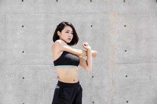 A healthy Japanese woman wearing black sportswear is stretching outdoors. Background is gray concrete.