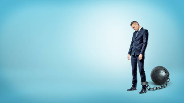 un uomo d'affari depresso su sfondo blu sta con una testa abbassata mentre è incatenato a una palla di ferro - debt trapped chain ball and chain foto e immagini stock