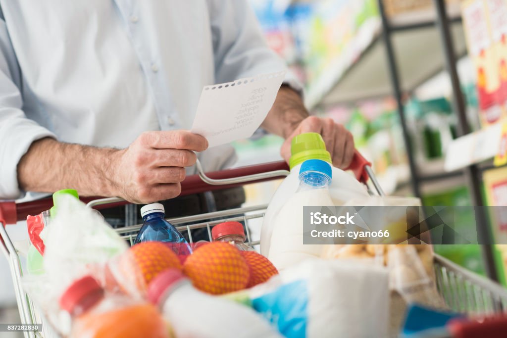 Man shopping with a grocery list Man at the supermarket shopping with a grocery list and pushing a full cart, lifestyle and retail concept Shopping Cart Stock Photo