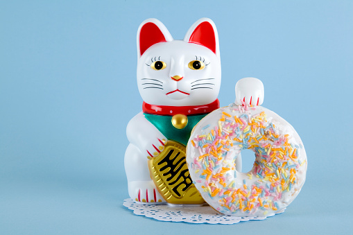 a maneki neko presenting a multicolor donuts on a doily paper and a pop colorful background.Minimal quirky color still life photography