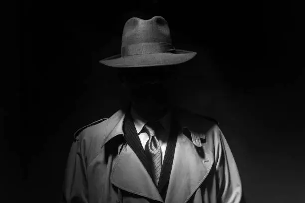 Man posing in the dark with a fedora hat and a trench coat, 1950s noir film style character