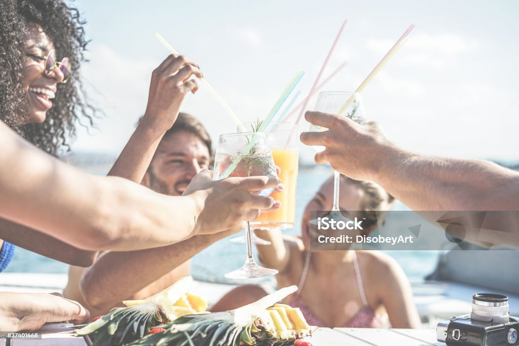 Happy friends drinking tropical cocktails at boat party and eating pineapple - Young people having fun in sea excursion - Youth and summer concept - Main focus on left bottom hand glass - Retro filter Friendship Stock Photo