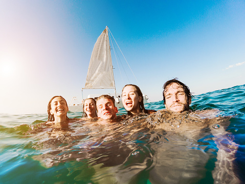 Happy friends swimming into the ocean next sailing catamaran boat - Young people having fun diving inside sea - Travel, tropical, youth, friendship and summer concept - Main focus on right guys