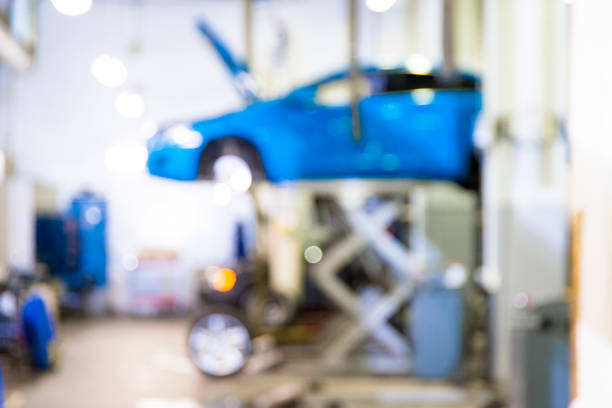 A white car with its hood open is being serviced outdoors. In the foreground, a large array of mechanic's tools is visible, including screwdrivers, pliers, and wrenches. A striped umbrella provides shade for the work area. The scene suggests a mobile mechanic service or roadside assistance, with all necessary equipment readily available for various car repairs.