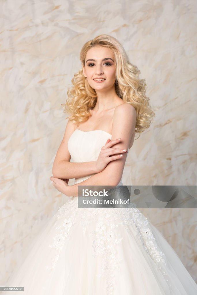 Fashionable White Gown Beautiful Blonde Model Bride Hairstyle And Makeup Concept Young Smiling Girl In Wedding Festive Dress Standing Indoors On Light Background Slender Woman Look Forward Stock Photo - Download Image Fashionable White Gown Beautiful Blonde Model Bride Hairstyle And Makeup Concept Young Smiling Girl In Wedding Festive Dress Standing Indoors On Light Background Slender Woman Look Forward Stock Photo - Download Image