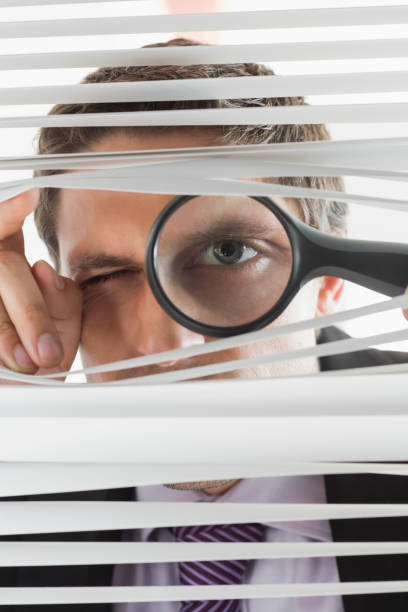 istock Businessman peeking through blinds with magnifying glass 837546364