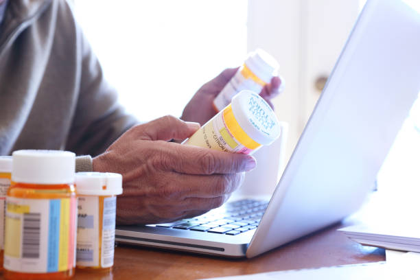 Close up Of Man Holding Prescription Medication Bottles In Front Of Laptop Close up of a Hispanic man in his late fifties holding two of his prescription medication bottles in his hands as he looks at his laptop computer while he sits at his dining room table.  Sunlight filters in through the window behind him bathing the room with a soft glow of light. prescription medicine stock pictures, royalty-free photos & images