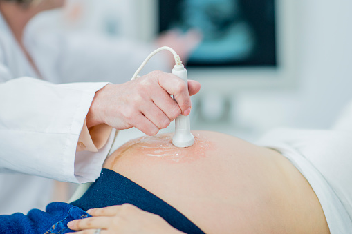 A pregnant asian woman is at the doctor's office getting a checkup to ensure the health of her unborn child. Here the doctor is using a doppler device to ultrasound the woman's stomach to see the baby. Just the stomach is visible in this frame.