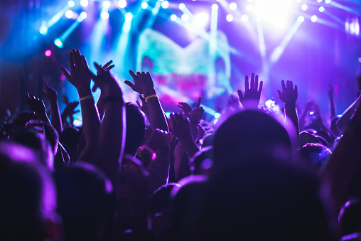 Rear view of a large group of people in front of a music festival stage. Crowd is excited and dancing, raising hands, clapping, punching the air, filming with mobile phones, etc...