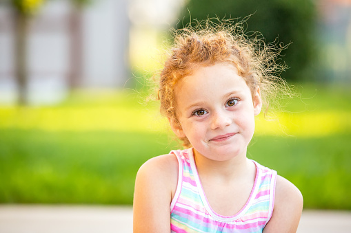 Close-up of a young girl with curly red hair pulled back in a ponytail. The girl is smiling a crooked smile and looking directly at the camera. Taken on a beautiful summer evening in the back yard.