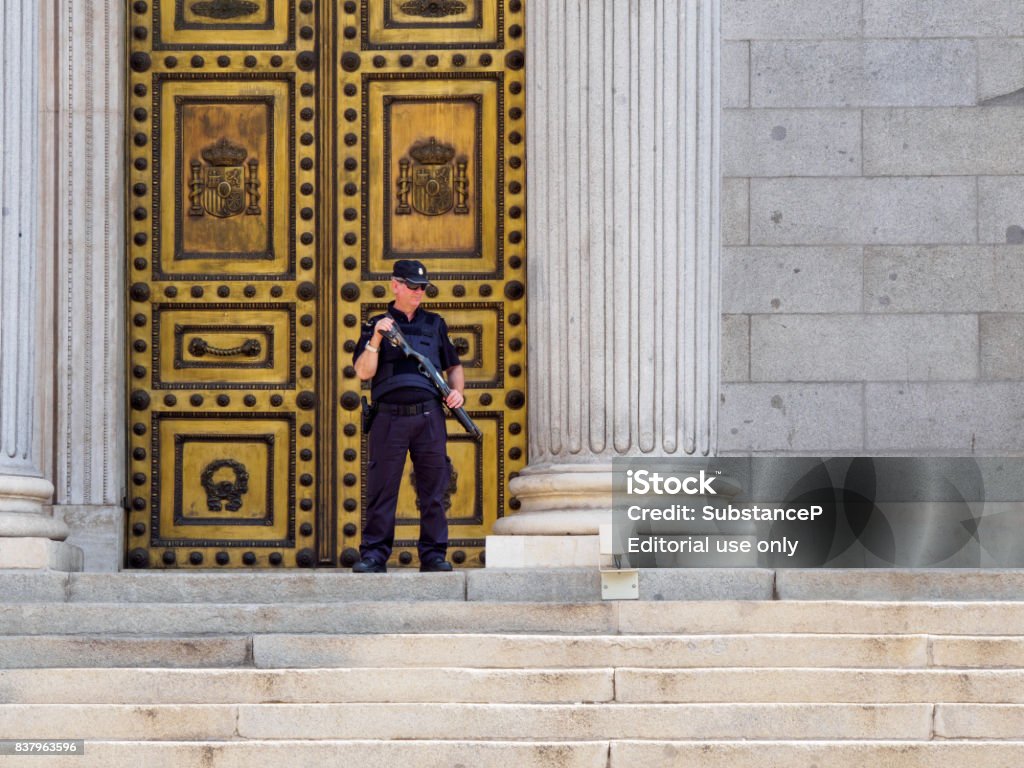 Armed policeman stands guard in front of Spanish Congress, Madrid, Spain June 21, 2017. A policeman armed with a shotgun and body armor stands guard in front of the doors of the Congress of Deputies, Madrid, Spain. Politics and security editorial concept. Adult Stock Photo