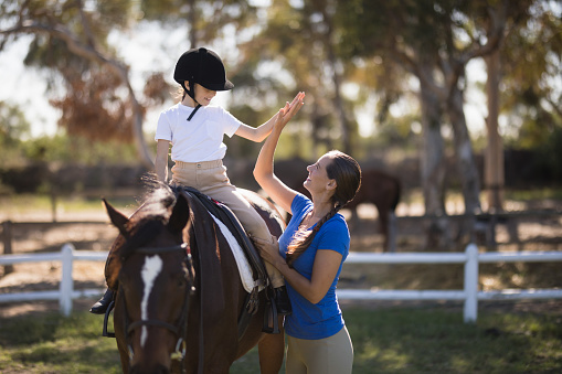 Side view of woman giving high five to girl sitting on horse in paddock