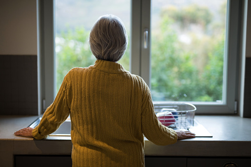 Rear view of senior woman standing near the kitchen sink and looking through window