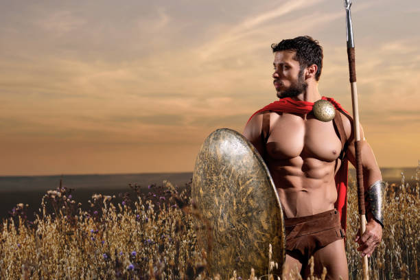 In his best shape. Horizontal shot of a muscular handsome young man Spartan warrior with perfect muscular strong body wearing a red cape with shield and spear standing alone in the field on dusk