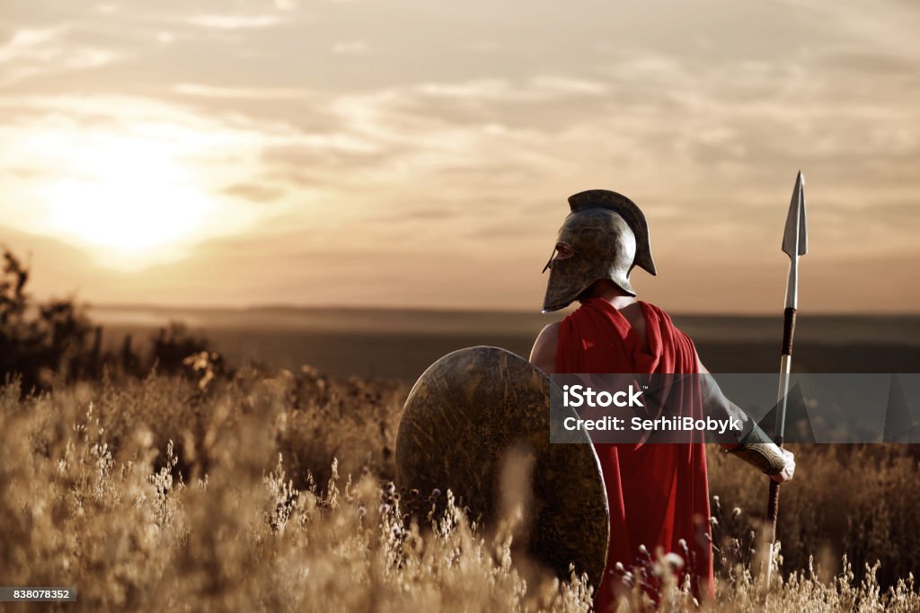 Warrior wearing iron helmet and red cloak. Back view of warrior man wearing iron helmet and red cloak holding sword, soldier looking and turned back. Male wearing like spartan or antique roman at field on sunset. Gladiator Stock Photo