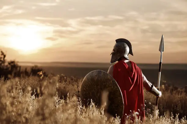 Back view of warrior man wearing iron helmet and red cloak holding sword, soldier looking and turned back. Male wearing like spartan or antique roman at field on sunset.