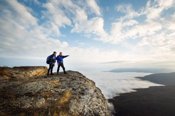 Photo of Two climbers standing on top of summit above clouds in the mountains. Hiker man pointing with his hand discussing route. Plan, vision and mission concept