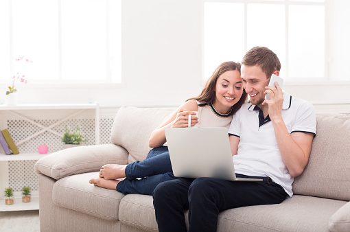 Young couple web surfing on laptop sitting on sofa at home, man talking on phone, relaxing, copy space