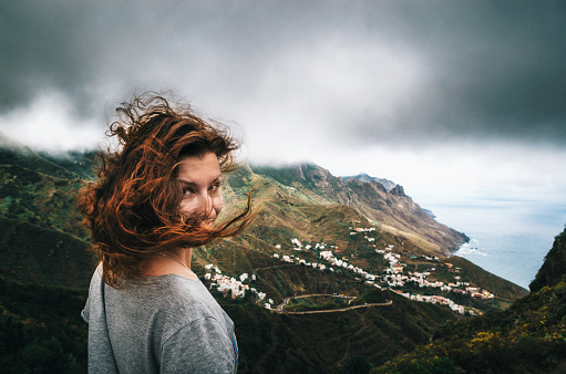 Joyful young woman with flying hair hidden her face looks at the camera in front of Taganana village on north of Tenerife. Concept of travelling, happiness, freedom. Traveler are enjoying the landscape, Canary Islands, Spain