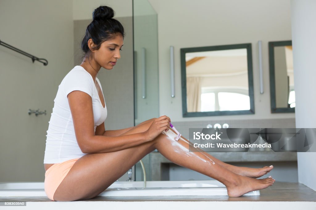 Full length of woman shaving leg in bathroom Full length of young woman shaving leg by bathtub in bathroom Shaving Stock Photo