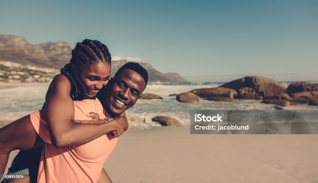 Young couple enjoying at the beach Young african man carrying his girlfriend on his back at the beach. Loving couple having piggyback fun on seaside. Couple - Relationship Stock Photo