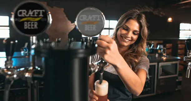 Beautiful young woman pouring beer into the glass. Female bartender tapping craft beer in bar.