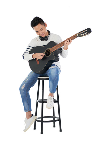 Talented smiling Asian man sitting on stool and playing guitar