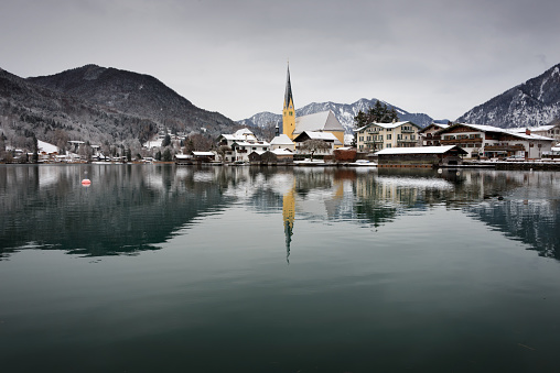 Church and houses in the village of Rottach-Egern at the edge of the water of Tegernsee lake with Alps mountains in the background in the snow in winter in Bavaria at the border between Germany and Austria.
