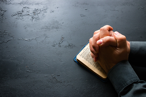 Hands of a young man folded praying over a Bible, hands over soft focus Bible, prayer and Bible study concept, contemplation concept