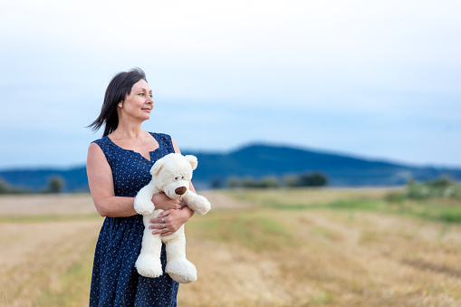 Portrait of senior woman with teddy bear at autumn wheat field. Concept of old peope dreaming about childhood. Countryside