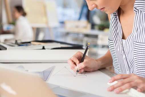 This is a closeup of an unrecognizable female architect as she leans in while sitting at her desk and concentrates on her drawing.  There is an incidental coworker working at her desk in the background.