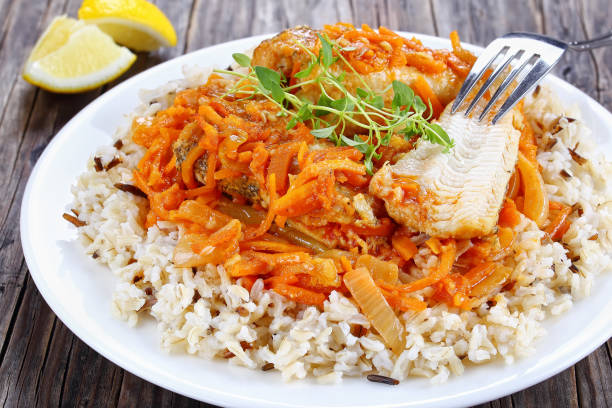 portion of white fish braised with carrot, onion, tomato sauce, spices and herbs with wild and long grain rice on plate on dark wooden table with lemon wedges on background, view from above, close-up