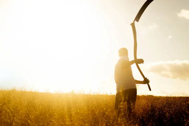 A young farmer rests with a scythe over his shoulders in his fields which are ripe with crops, on a sunny evening.