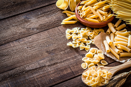High angle view of a rustic wooden table with a large Italian pasta variety arranged at the right and leaving useful copy space for text and/or logo. The types of pasta included are spaghetti, orecchiette, conchiglie, rigatoni, fusilli, penne and tagliatelle. . Predominant colors are yellow and brown. DSRL studio photo taken with Canon EOS 5D Mk II and Canon EF 100mm f/2.8L Macro IS USM