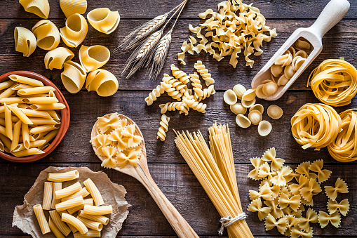 Top view of a rustic wooden table filled with a large Italian pasta variety. The types of pasta included are spaghetti, orecchiette, conchiglie, rigatoni, fusilli, penne and tagliatelle. Predominant colors are yellow and brown. DSRL studio photo taken with Canon EOS 5D Mk II and Canon EF 100mm f/2.8L Macro IS USM