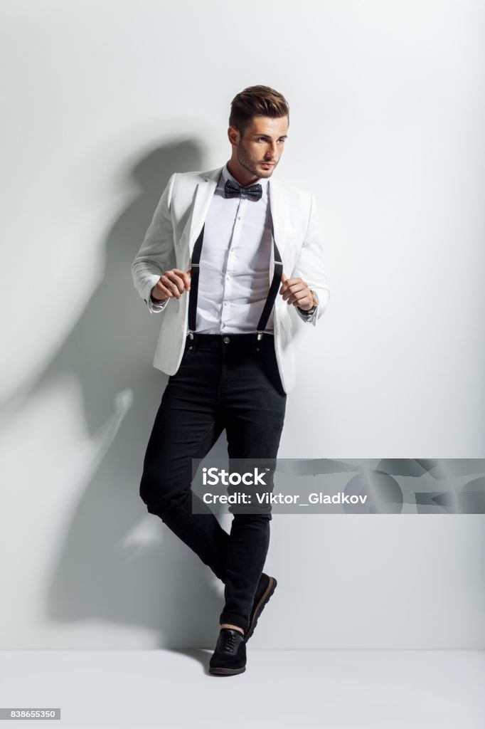 Full length portrait of handsome stylish man in white elegant suit posing and leaning at wall Tuxedo Stock Photo