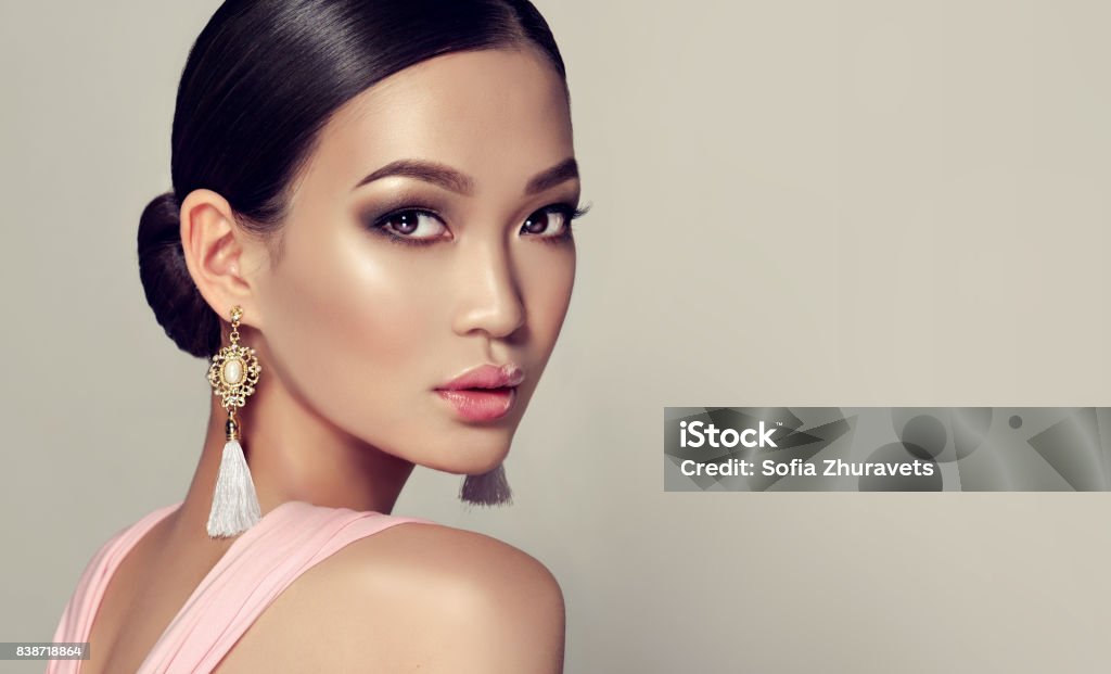 Young, gorgeous asian woman in a smoky eyes style make up and tassel earrings Young, gorgeous asian fashion-model put on in a smoky eyes style make up, black hair gattered in a beam. dressed in tassel earrings and  pink gown. Asian beauty. Women Stock Photo