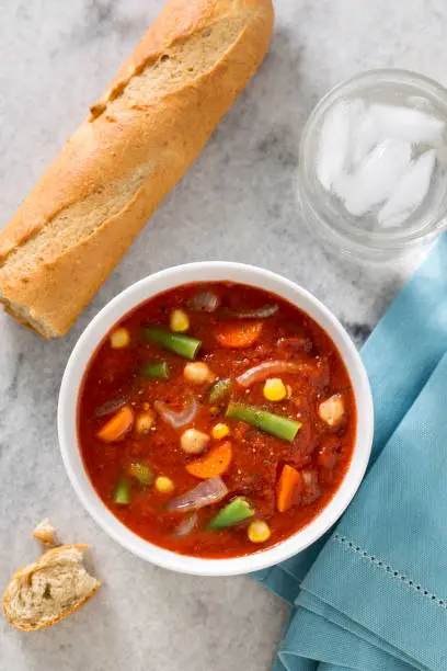 Bowl of hearty vegetable soup with rustic loaf of bread on white marble counter top.
