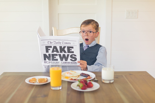 A young buisnessman dressed in a business suit is sitting at the dining room table with a great American breakfast of eggs, bacon, toast and orange juice while he is reading the shocking version of fake news. Can the media be trusted anymore?