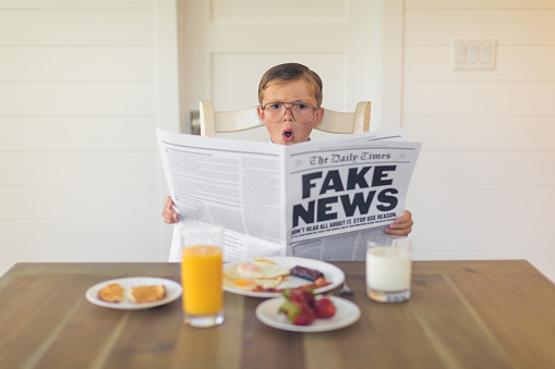 A young buisnessman dressed in a business suit is sitting at the dining room table with a great American breakfast of eggs, bacon, toast and orange juice while he is reading the shocking version of fake news. Can the media be trusted anymore? Journalism is off the rails.