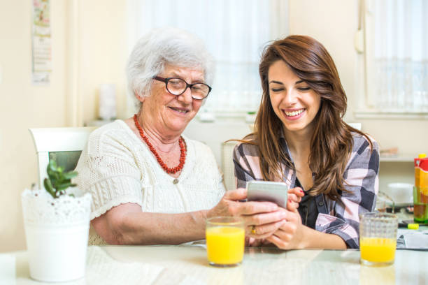 grandmother and her granddaughter using phone while sitting near the table together at home. - wireless technology cheerful granddaughter grandmother imagens e fotografias de stock