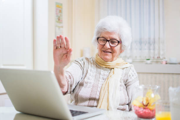 mujer senior canas agitando la mano en frente del ordenador portátil teniendo video llamada con miembros de su familia. - one person lifestyles 80 plus years indoors fotografías e imágenes de stock