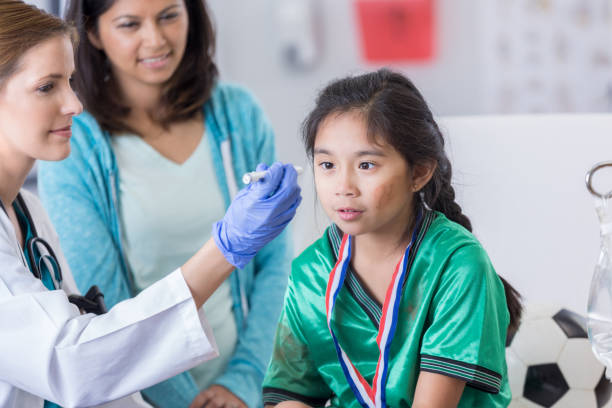 Emergency room doctor examines young dazed soccer player Female emergency room doctor uses a pen light to examine a young soccer player's eyes. The girl has a dazed expression on her face. She is wearing a soccer uniform. concussion stock pictures, royalty-free photos & images