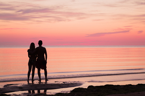 Young couple looking far away at the seaside in summer evening. Man's and woman's silhouettes on the beach. Sea water reflecting a pink heavenly colors and creating a romantic atmosphere.