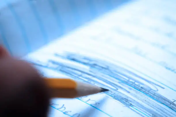 A close up of  someone holding a pencil as they scribble out something that had been previously written into a notebook.