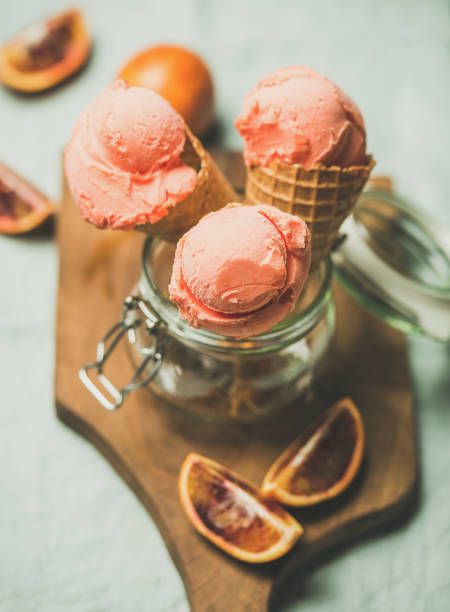 Refreshing summer blood orange ice cream or sorbet scoops in sweet waffle cones in glass jar served with fresh orange slices on wooden board over light grey towel background, selective focus